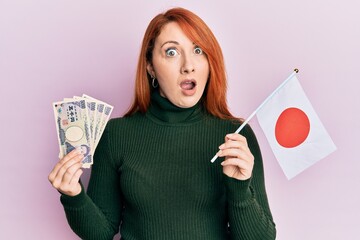 Sticker - Beautiful redhead woman holding 5000 japanese yen banknotes and japan flag in shock face, looking skeptical and sarcastic, surprised with open mouth