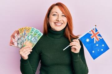 Canvas Print - Beautiful redhead woman holding australian dollars and australia flag smiling with a happy and cool smile on face. showing teeth.