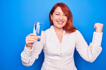 Sticker - Young beautiful redhead woman drinking glass of water over isolated blue background screaming proud, celebrating victory and success very excited with raised arm