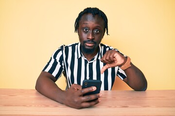 Poster - Young african american man with braids using smartphone sitting on the table with angry face, negative sign showing dislike with thumbs down, rejection concept