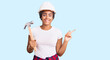 © Krakenimages.com - Young african american woman with braids wearing hardhat holding hammer smiling happy pointing with hand and finger to the side