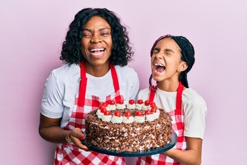 Poster - Beautiful african american mother and daughter wearing baker apron holding homemade cake smiling and laughing hard out loud because funny crazy joke.