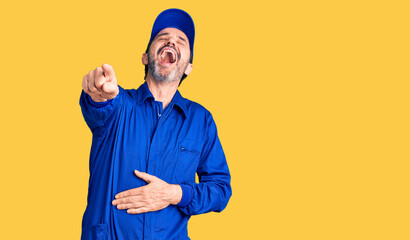 Middle age handsome man wearing mechanic uniform laughing at you, pointing finger to the camera with hand over body, shame expression