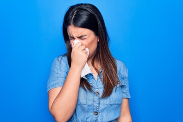 Sticker - Young beautiful woman sick for flu. Using paper handkerchief on full nose of mucus standing over isolated blue background