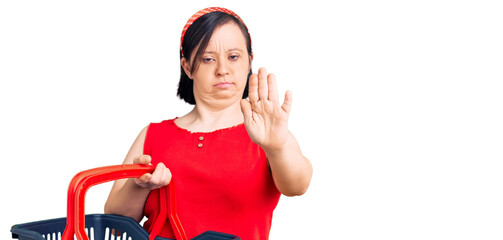 Wall Mural - Brunette woman with down syndrome holding supermarket shopping basket with open hand doing stop sign with serious and confident expression, defense gesture