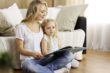 Happy family. Blonde young mother reading a book to her cute daughter while sitting at wooden floor. Motherhood concept