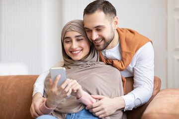 Wall Mural - Happy Muslim Couple Using Smartphone Texting Sitting On Couch Indoors