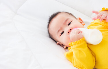 Asian beautiful little baby girl wearing a yellow dress eat milk sleeping feeding lying on the white bed, infant holding a bottle of milk, baby food concept