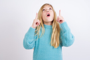 Wall Mural - Cute Caucasian kid girl wearing blue knitted sweater against white wall being amazed and surprised looking and pointing up with fingers showing something strange.