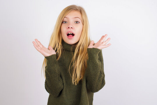 Surprised terrified Caucasian kid girl wearing green knitted sweater  Gestures with uncertainty, stares at camera, puzzled as doesn't know answer on tricky question, People, body language, 