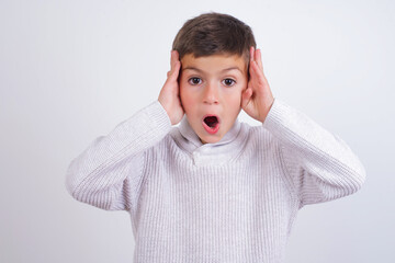 Wall Mural - Cute Caucasian kid boy wearing knitted sweater against white wall with scared expression, keeps hands on head, jaw dropped, has terrific expression. Omg concept