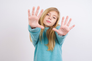 Wall Mural - Portrait of smiling Cute Caucasian kid girl wearing blue knitted sweater against white wall looking at camera and gesturing finger frame. Creativity and photography concept.