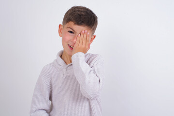 Wall Mural - Cute Caucasian kid boy wearing knitted sweater against white wall covering one eye with her hand, confident smile on face and surprise emotion.