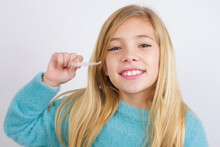Cute Caucasian Kid Girl Wearing Blue Knitted Sweater Against White Wall Holding Invisible Aligner Braces. Recommendation And Dental Healthcare Concept.