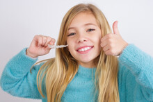 Cute Caucasian Kid Girl Wearing Blue Knitted Sweater Against White Wall Holding Invisible Aligner Braces And Doing Ok Sign. Recommendation And Dental Healthcare Concept.