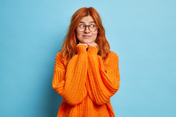 Wall Mural - Pretty young woman with natural red hair keeps hands under chin and looks aside thinks about something pleasant wears optical glasses and knitted orange jumper isolated over blue background.