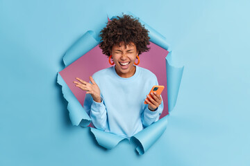 Wall Mural - Overjoyed dark skinned woman with curly hair raises palm and laughs out joyfully holds modern mobile phone in hand uses modern device for texting or messaging breaks through paper background