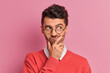 © WHstudio Leushin N - Handsome thoughtful man holds chin and looks doubtful away purses lips makes planning tries to decide what to do better has stylish hairdo wears transparent round glasses poses indoor over rosy wall
