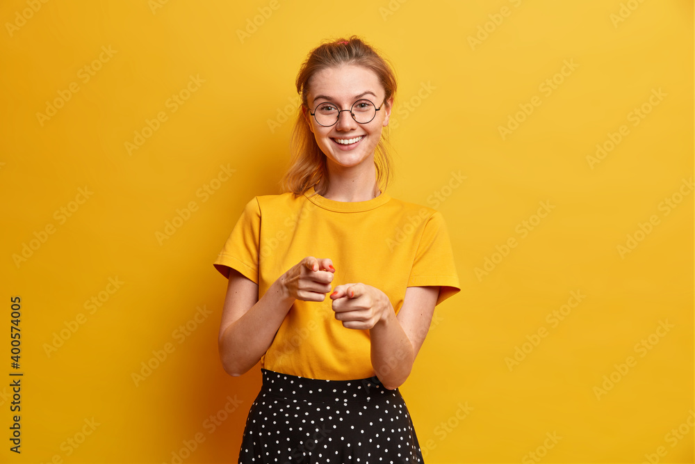 Pretty European girl with glad expression points index fingers directly at camera selects you smiles friendly wears casual t shirt and polka dot skirt isolated over yellow background. I choose this - obrazy, fototapety, plakaty 