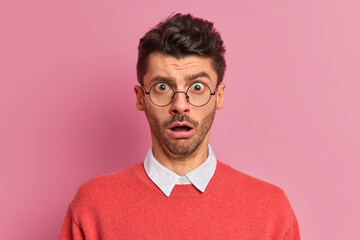 Wall Mural - Close up shot of amazed shocked man holds breath from shock stares through optical glasses opens mouth hears surprising news dressed in white shirt with jumper isolated over pink background.