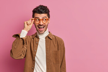 Horizontal shot of happy European man with bristle keeps hand on spectacles and smiles positively concentrated aside on copy space rejoices nice offer dressed casually poses against pink background