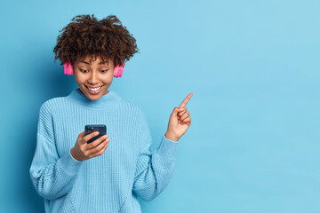 Wall Mural - Photo of beautiful dark skinned curly woman concentrated in smartphone chooses song to listen enjoys favorite music via headphones wears blue jumper indicates on blank space over blue background