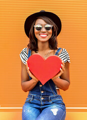 Beautiful smiling african woman holding a red heart. Fashion portrait stylish pretty woman in sunglasses outdoor.