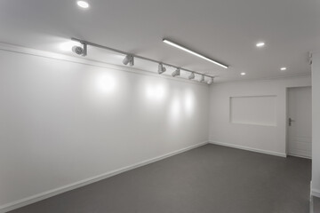 Empty white wall without frames in gallery room. Illuminated space for art exposition. Gallery lighting, spotlights No people. Showroom with a dark gray self-leveling floor.