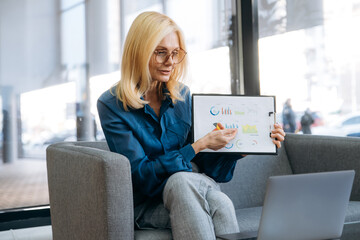 Elegant senior lady in eyeglasses talk with colleagues by video call, show them financial graphs, discuss about project. Beautiful business woman sit at the couch in formal wear, distant work concept