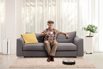 Poster - Robotic vacuum cleaner cleaning the floor and an elderly man drinking tea on a sofa