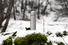 Steel Vacuum Thermos Outside In The Winter Forest. Traveling Aluminum Flask Standing On Moss Covered With Snow.