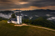 Jamnik, Slovenia - Aerial drone view of golden sumer sunrise at Jamnik St.Primoz church. Morning fog gently goes in the mountains behind the chapel with colorful sky and Julian Alps at background
