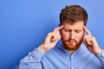 portrait of thoughtful caucasian guy thinking isolated over blue background, stand with eyes closed, holding head, concentrated on thoughts