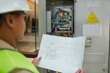 Female worker holding blueprints while inspecting electrical switchboard at construction site, copy space