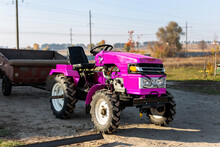 Small Mini Glamour Modern Pink New Tractor With Trailer Standing Near Hangar Building Farm Countryside During Sunset Or Sunrise. Small Agricultural Machinery. Rural Country Farmland Scene Background