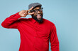 Excited funny young bearded african american man 20s wearing casual red shirt cap eyeglasses standing showing victory sign looking camera isolated on pastel blue color background studio portrait.