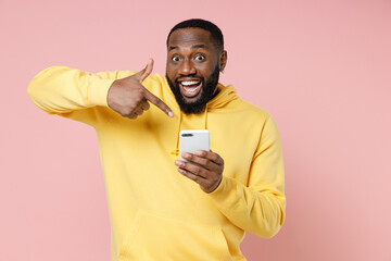 Wall Mural - Excited young african american man 20s wearing casual yellow streetwear hoodie pointing index finger on mobile cell phone typing sms message isolated on pastel pink color background studio portrait.
