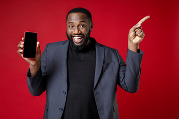 Funny young african american business man 20s in classic jacket suit standing hold mobile phone with blank empty screen pointing index finger aside up isolated on red color background studio portrait.