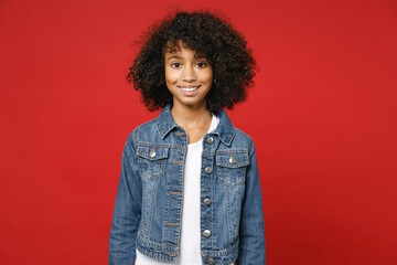 Smiling funny little african american kid girl 12-13 years old wearing casual denim jacket looking camera isolated on bright red color background children studio portrait. Childhood lifestyle concept.