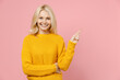 © ViDi Studio - Smiling elderly gray-haired blonde woman lady 40s 50s years old in yellow casual sweater pointing index finger aside up on mock up copy space isolated on pastel pink color background studio portrait.