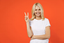 Smiling Funny Elderly Gray-haired Blonde Woman Lady 40s 50s Years Old In White Casual T-shirt Standing Showing Victory Sign Looking Camera Isolated On Bright Orange Color Background Studio Portrait.