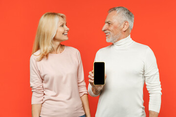 Funny couple two friends elderly gray-haired man blonde woman in white pink clothes hold mobile cell phone with blank empty screen looking at each other isolated on orange background studio portrait.