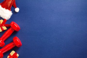 Wall Mural - Exercise, fitness and developing the concept of a merry Christmas and a happy New year. Red dumbbells, Santa hat and gifts with red ribbons on a dark blue background. Flat lay, top view