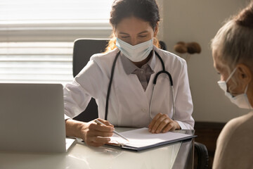 Young female GP or therapist in medical facial mask consult mature patient about health insurance in hospital. Woman doctor in facemask consider results or treatment with elderly client in clinic.