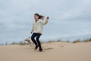 Wall Mural - cute young girl running in the dunes with her dog in autumn