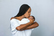 © Ursula Page - A beautiful young African-American woman with braids is kissing her newborn son and looking at him with love on a white gray background and giving him a kiss