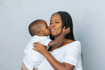 A beautiful young African-American woman with braids is holding her toddler age cute son and he is kissing her with a white gray background.