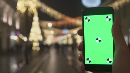 Portrait young woman hand holding a mobile phone with a vertical green screen in night street on Christmas time. Magic atmospheric city light. Slow motion