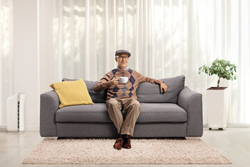 Poster - Elderly man seated on a sofa holding a cup and smiling