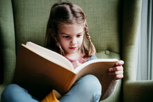 Girl Reading Book While Sitting On Chair At Home